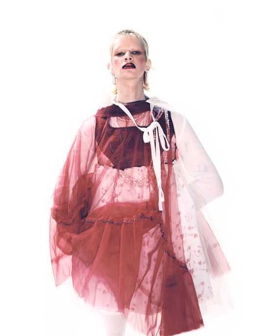 A high-fashion, editorial image of a person wearing a pinky-red, see-through dress made of net paired with vegan pearl jewellery from Vellva