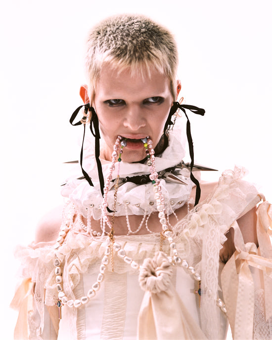 Close up of a person with short, cropped, bleach blonde hair with a high-fashion outfit featuring a historical-style soft pink collar biting a strand of Vellva vegan pearls which is also draped around their neck