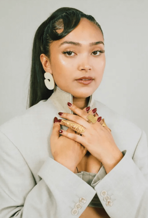 A close up of singer Joy Crookes wearing a soft grey suit jacket paired with statement grey earrings and deep red nails. She holds her hands up against her neck, hiding her Vellva vegan pearl necklace