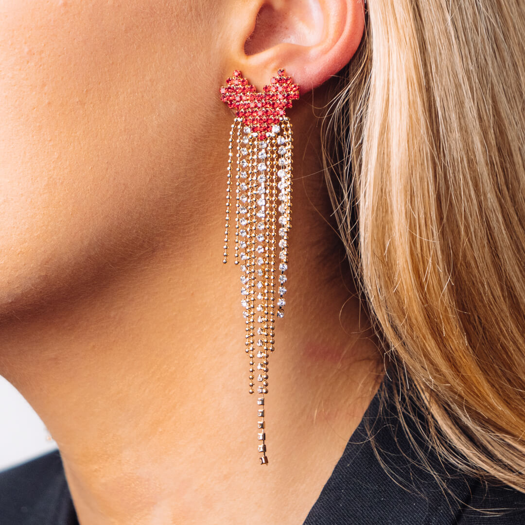 A pair of large earrings made up of a red heart with a gold tone and diamante tail worn on blonde woman.
