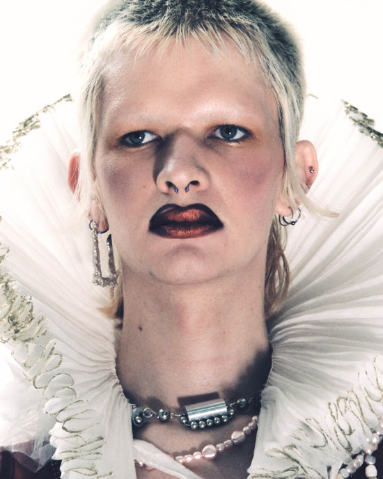 A very close up image of a person with cropped, bleached blonde hair, bleached blonde eyebrows and dramatic metallic red lipstick wearing a dramatic collar in the style of Tudor royalty and a pink handmade Vellva vegan pearl necklace  
