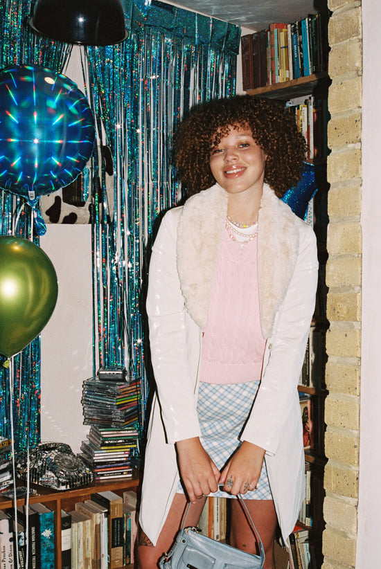A happy teenage girl stands in front of party decorations wearing a soft pink faux fur coat, a blue checked skirt, soft pink jumper and three vegan pearl necklaces in soft pastel colours 