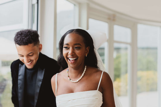 A smiling bride stands next to her groom wearing a white dress with spaghetti straps and Vellva's Il Mare Necklace - a handmade vegan pearl and beaded necklace