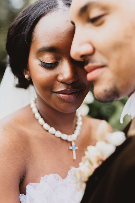 A very close up image of the smiling faces of a newly married couple focussing on the bride who is wearing a bandeau-style white wedding dress and Vellva's La Contessa Necklace, a chunky vegan pearl necklace with a pink cross pendant