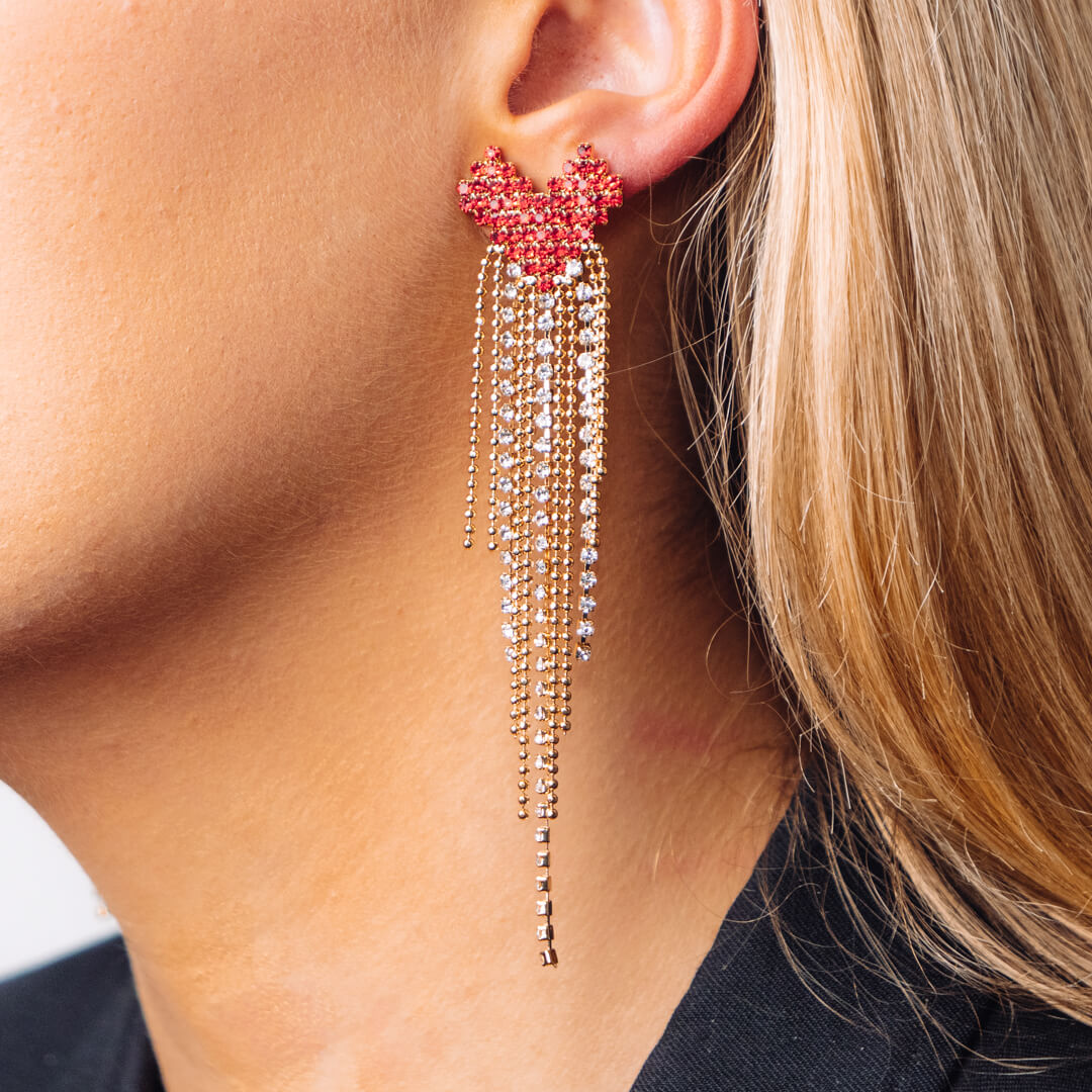 A pair of large earrings made up of a red heart with a gold tone and diamante tail worn on blonde woman. 