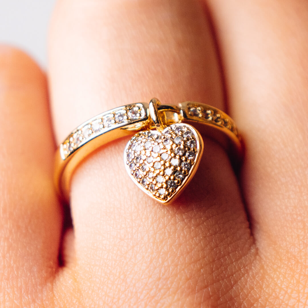 A gold tone and diamante ring with a heart shaped pendant worn on a finger.