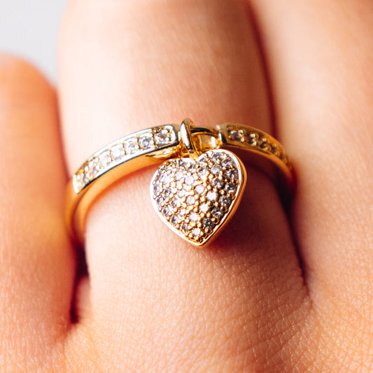 A gold tone and diamante ring with a heart shaped pendant worn on a finger.