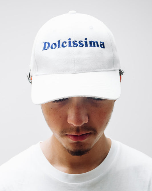 A front on view of a man wearing a white cotton slogan cap with the Italian word Dolcissima embroidered on it in dark blue.